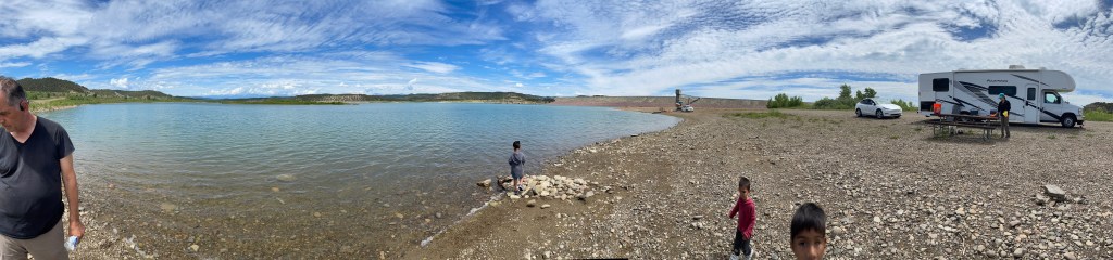 Panoramic view of a peaceful lake scene with a recreational vehicle parked nearby, kids playing on the rocky shore, and a blue sky filled with clouds.