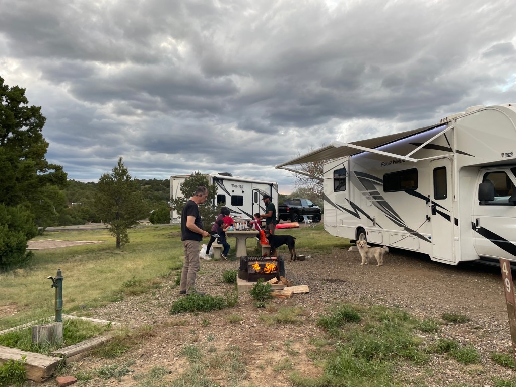 A family gathers around a campfire near their RV, with two dogs present. The sky is cloudy, and they appear to be enjoying an outdoor moment together.