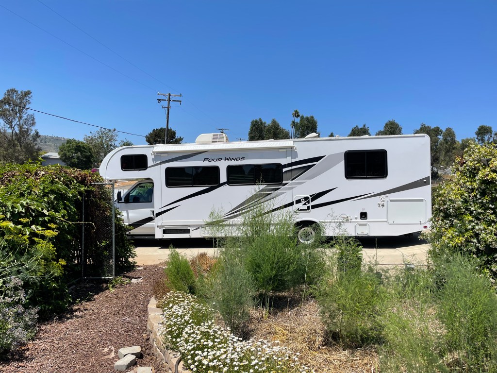 A mid-size Class C RV parked in a residential area, surrounded by plants and greenery under a clear blue sky.
