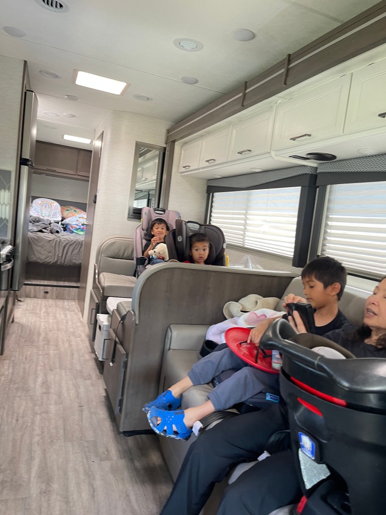 Interior view of a mid-size Class C RV with three young children in car seats and a woman sitting nearby, showcasing a family-friendly layout.